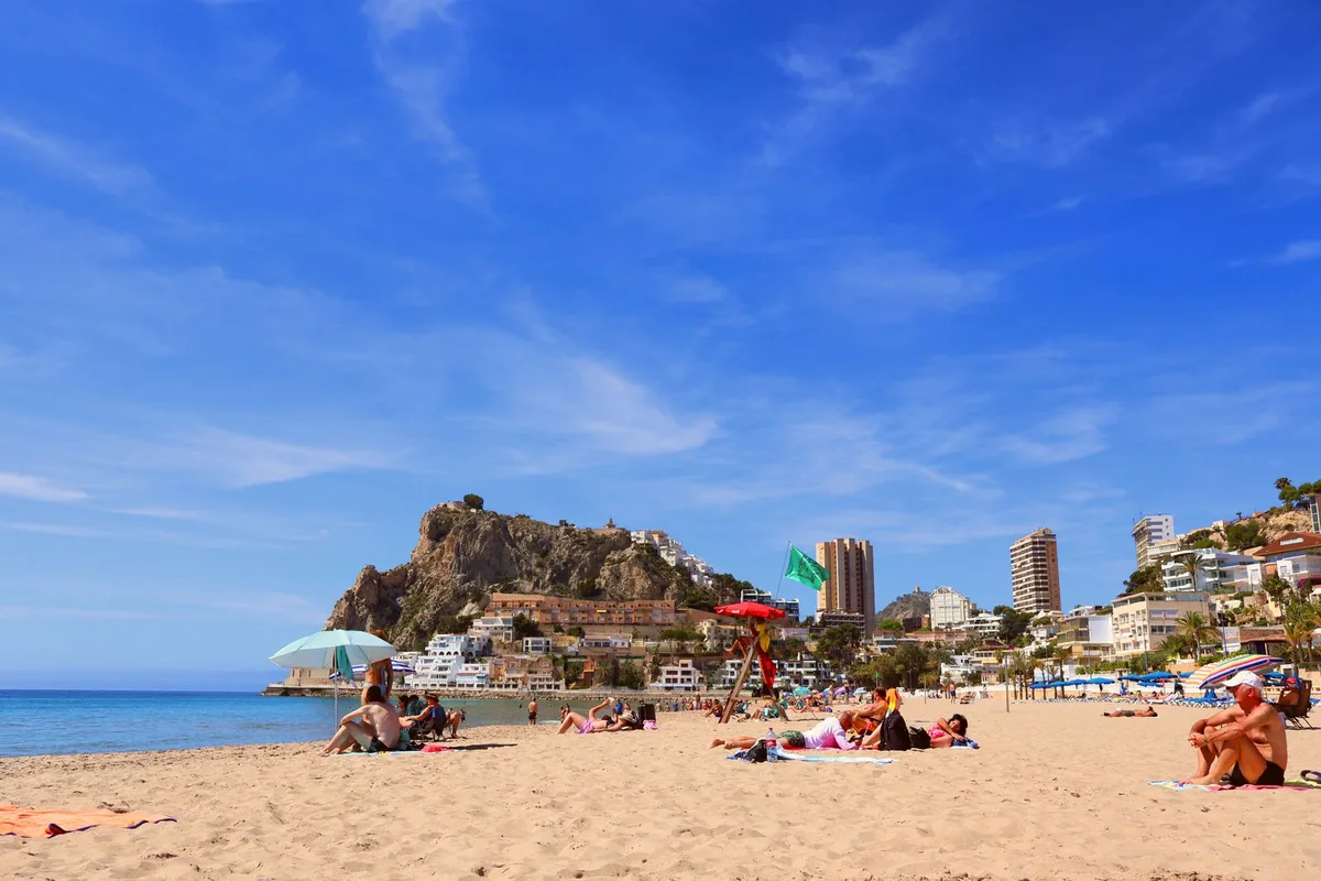 A sunny beach with people sunbathing under umbrellas, a lifeguard post, and a rocky headland and tall buildings behind (Alicante).