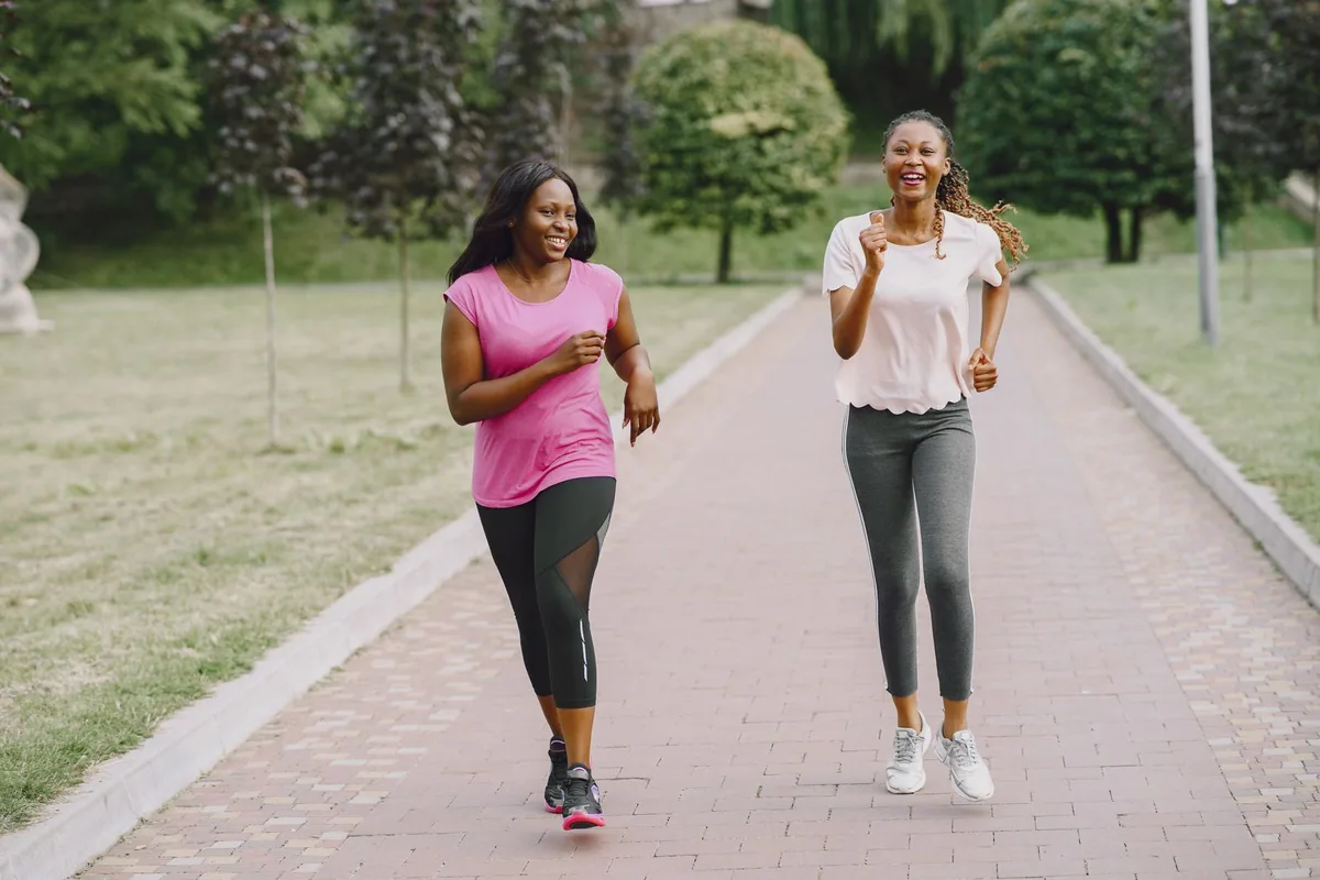 Two young women in pink T-shirts jogging and laughing along a path in a green park.