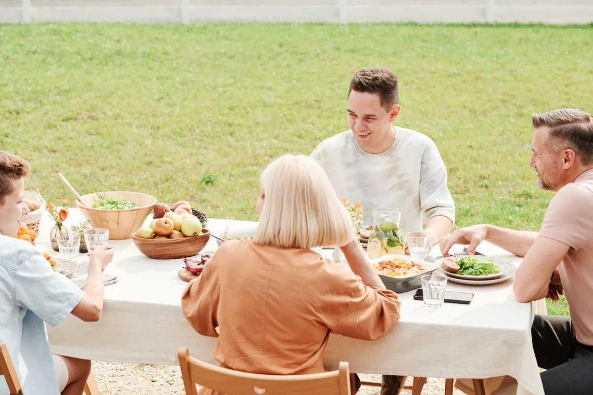 A family of four having lunch outdoors at a long table with a white cloth, with salad, pasta, fruit and a jug of lemonade.