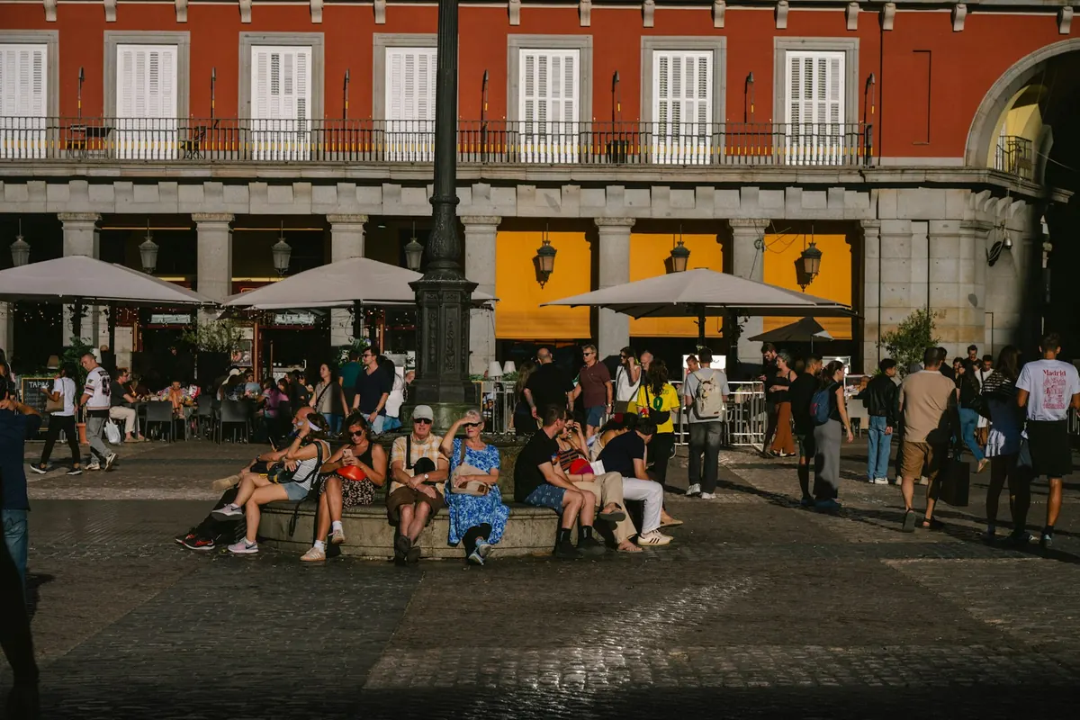 A sunny square with red buildings and stone arcades: people sit round a lamp-post in the middle and café terraces with big umbrellas fill the background (Plaza Mayor, Madrid).