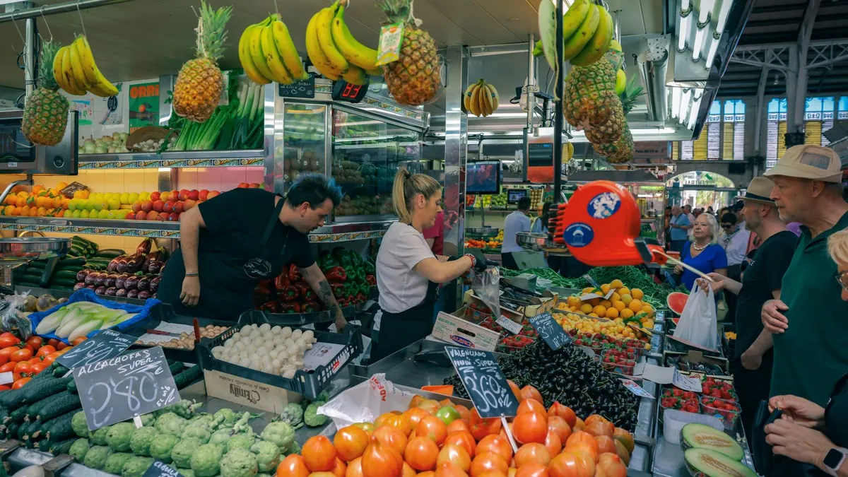A busy fruit and vegetable stall in an indoor market: two stallholders in black aprons serve older shoppers among tomatoes, cherries and melons (Mercado Central, Valencia).