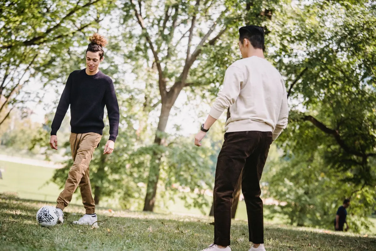 Two young men playing football on the grass under big leafy trees in a sunny park.