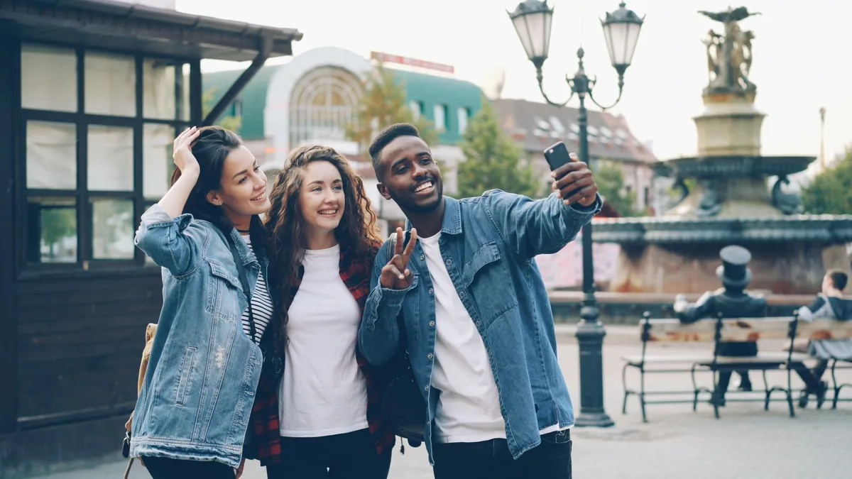Three smiling friends taking a selfie with a phone in a town square, with a fountain and street lamps behind.
