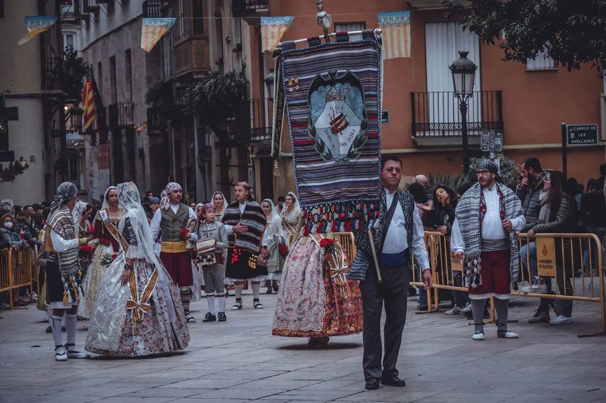 A festival procession in an old street in Valencia: women in long, richly embroidered traditional dresses with lace veils, men in traditional costume with striped blankets over their shoulders, and a man in front carrying a large embroidered banner. People watch from behind barriers and bunting hangs across the street (Las Fallas).
