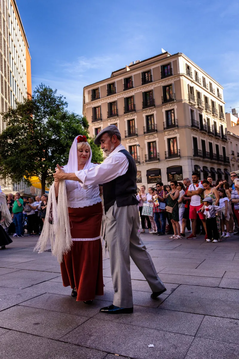 An older couple in traditional Madrid dress dancing in a sunny square: she wears a white headscarf with red carnations, a fringed white shawl and a long red skirt; he wears a flat cap, a white shirt and a dark waistcoat. A crowd behind them watches and films with their phones (San Isidro festival).