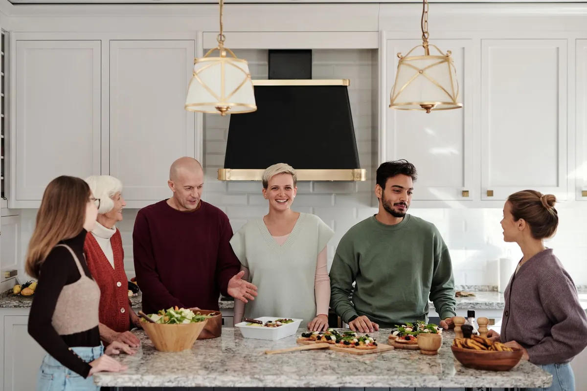 Six people of different ages, four women and two men, standing round a kitchen island (a table) in a white kitchen, chatting and smiling. On the worktop there is a big bowl of salad, small pizzas on wooden boards and a bowl of bread.