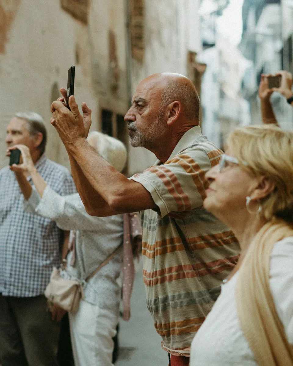 Older tourists in a narrow old street in Spain taking photos with their mobile phones; a smiling woman with glasses is in the foreground.