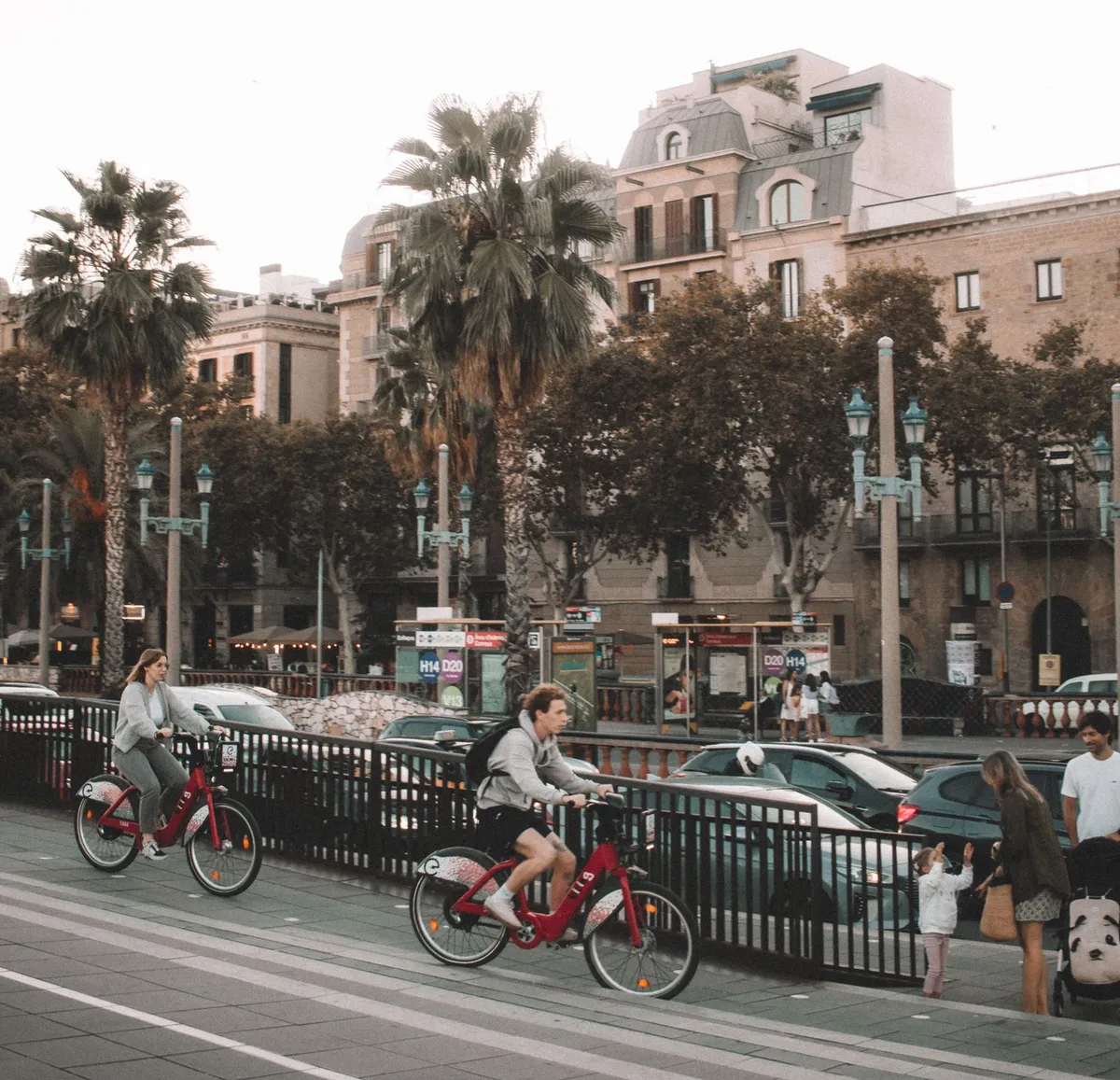A young woman and a young man riding red bicycles along a cycle path in Barcelona, with palm trees and old buildings behind, cars on the road and, on the right, a woman with a small child and a pushchair.